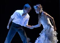 Ein Mann in weißem Hemd und eine Frau im weißen Kleid mit Blumenkranz halten sich auf einer Bühne an den Händen und blicken sich intensiv an.