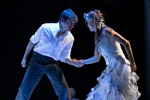 Ein Mann in weißem Hemd und eine Frau im weißen Kleid mit Blumenkranz halten sich auf einer Bühne an den Händen und blicken sich intensiv an.