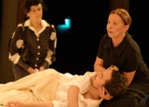 Eine Frau kniet neben einem Mann in weißer Kleidung, der auf dem Boden liegt. Im Hintergrund kniet eine weitere Frau in einem gepunkteten Kostüm.