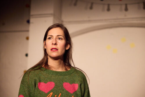 Eine Frau im grünen Pullover mit rosafarbenen Herzen und roten Lippen blickt nach oben.