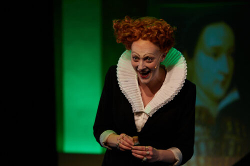 Eine Frau mit roten, hochgesteckten Haaren und weißem Rüschenkragen hält einen kleinen Behälter und lächelt.