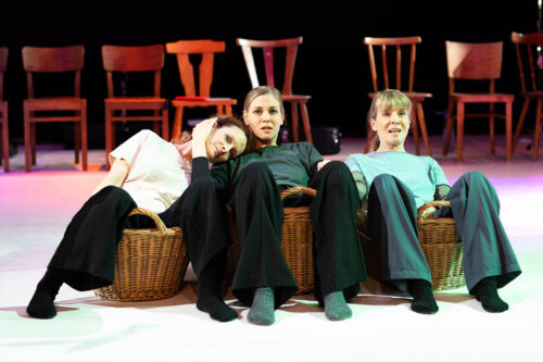 Drei Frauen sitzen entspannt in Weidenkörben auf einer Bühne. Eine lehnt ihren Kopf auf die Schulter der Nachbarin.