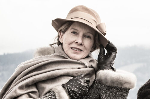 Freundliche Frau mit Hut, Handschuhen und warmem Überwurf lächelt in die Kamera.