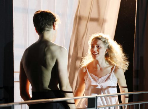 Eine lachende blonde Frau in einem weißen, trägerlosen Oberteil steht auf einer Balustrade und blickt einen Mann mit nacktem Oberkörper an, dessen Rücken zum Betrachter gewandt ist.