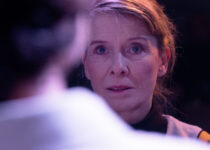 Nahaufnahme einer Frau auf der Bühne, die direkt in die Kamera schaut, mit einem unscharfen Rücken im Vordergrund. Violettes Licht.