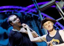 Mann in Brille und Schwarz hält ein Seil, der andere mit Piratenhut und Trägershirt hält es fest. Lila beleuchteter Hintergrund.