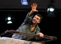 Ein lächelnder Schauspieler lehnt über ein Bettgestell und streckt die Hand hoffnungsvoll nach oben. Scheinwerfer beleuchten die Bühne.